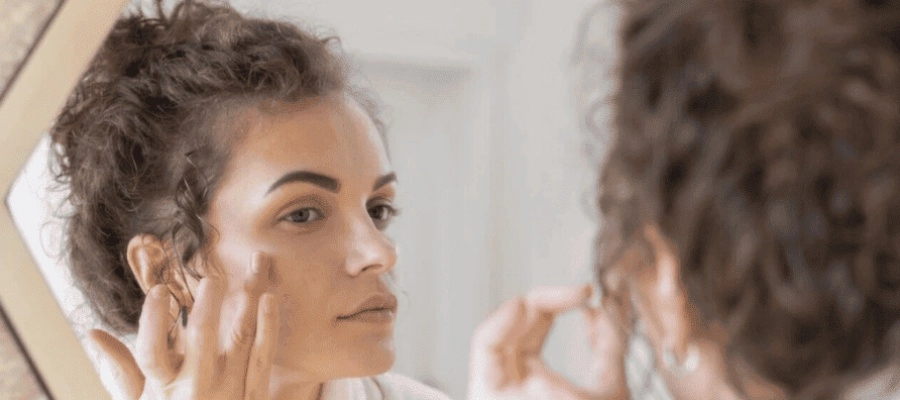Mulher com cabelos cacheados aplica creme no rosto enquanto se olha no espelho, em ambiente claro.
