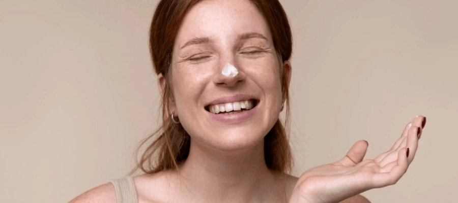 Mulher sorri com creme no nariz e olhos fechados em fundo bege.