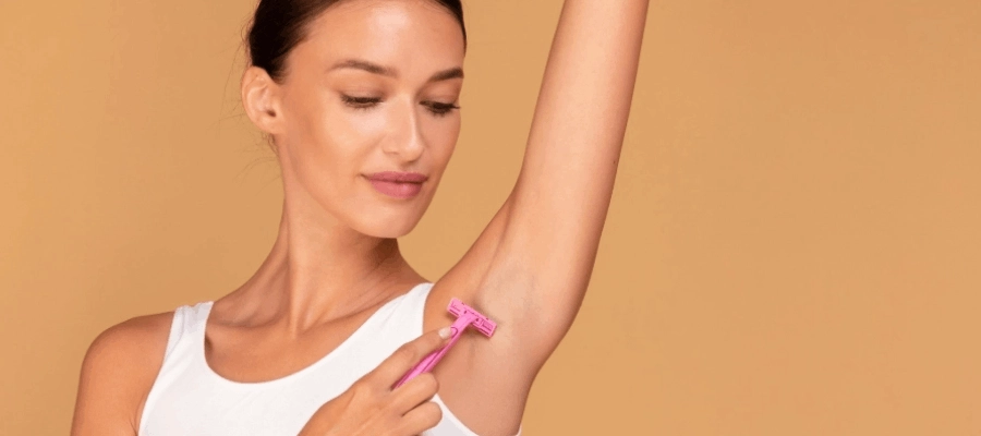 Mulher veste uma blusa branca e depila a axila com lâmina rosa.