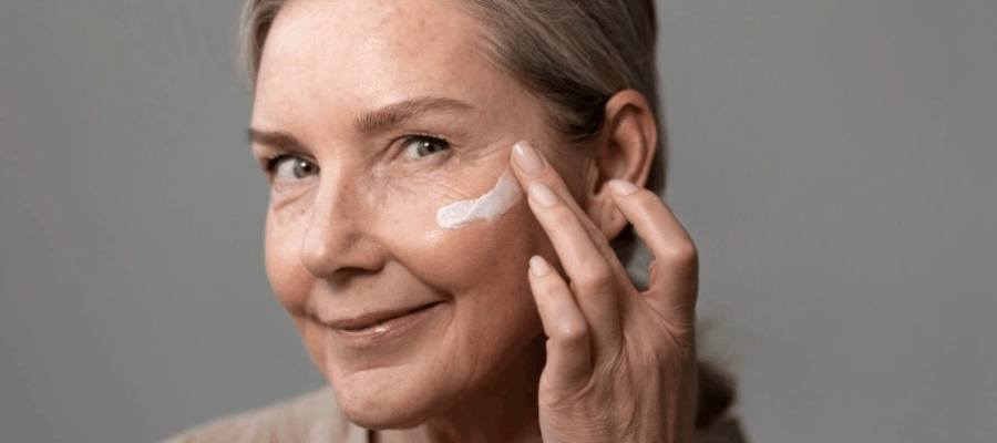 Mulher com pele madura sorri e aplica protetor solar no rosto.