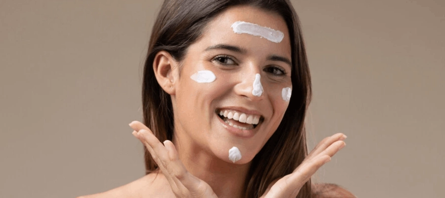 mulher jovem com cabelos longos sorridente aplicando protetor solar no rosto