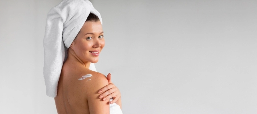 Mulher sorri com toalha na cabeça e aplica hidratante corporal com ceramida no ombro