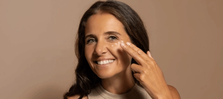 Mulher sorridente aplica creme na região dos olhos e evidencia como diminuir olheiras com skincare