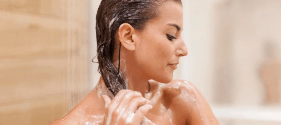 Mulher lava o cabelo com shampoo durante o banho, com espumas ao redor.